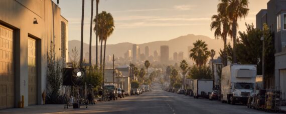 Los Angeles production street with studio lighting, palm trees, trailers, and city skyline at golden hour.